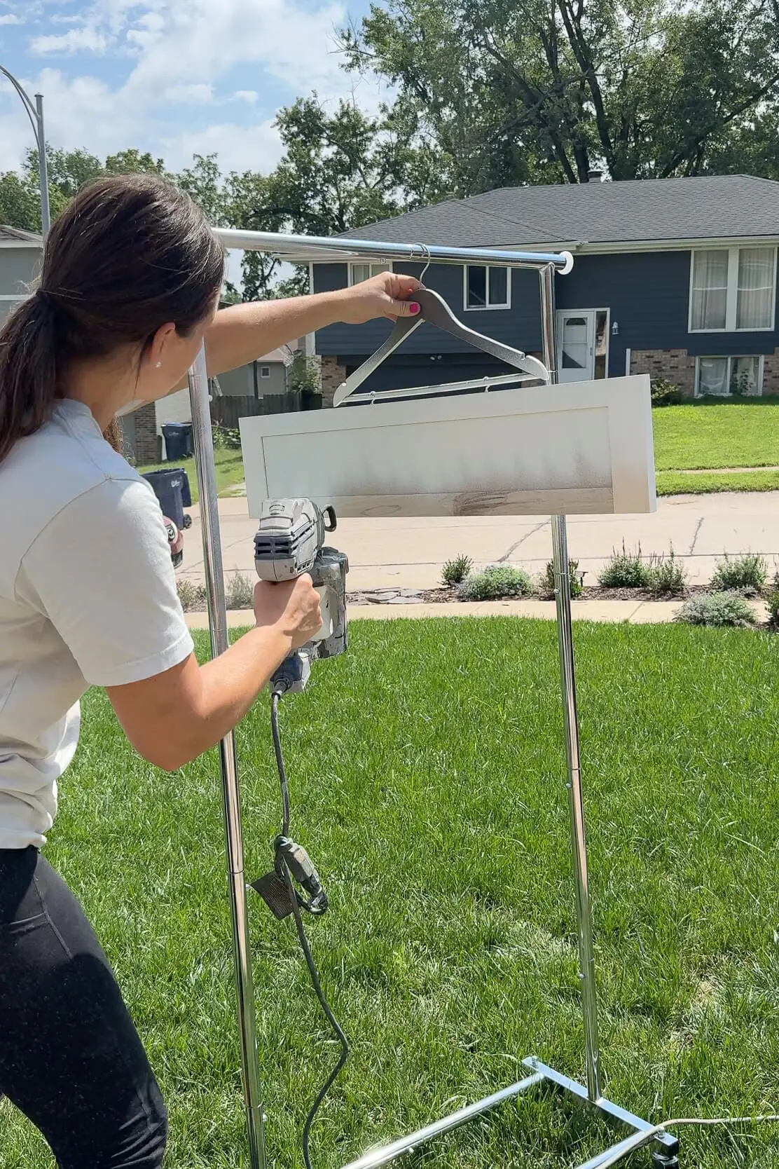 Person using a paint sprayer outdoors to paint a wooden cabinet door frame hanging from a clothing rack in a sunny yard with a suburban neighborhood in the background.